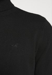 Svart turtleneck-tröja tillverkad av ribbad stickad tyg. Har en liten, tonad logotyp på bröstet och visar en figurnära design.