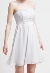 Robe blanche sans bretelles avec un corsage ajusté ornée de détails en dentelle et une jupe évasée. Confectionnée en tissu léger et lisse.