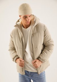 Veste matelassée en velours côtelé beige avec capuche, doublée de matière douce. Poignets et ourlet côtelés avec fermeture éclair frontale ; associée à une chemise blanche.