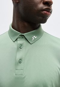 Άνδρας που φοράει ανοιχτοπράσινο πόλο με τρία κουμπιά και λευκό κεντημένο λογότυπο στο γιακά, κοιτώντας ελαφρώς προς τα δεξιά.