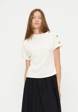 Femme aux cheveux bruns raides portant un haut blanc à manches courtes avec des boutons dorés sur l'épaule et une jupe noire sur fond blanc.