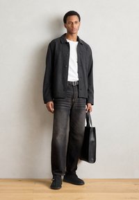 Chemise noire à boutons, t-shirt blanc, jeans droits noirs délavés, chaussures noires à enfiler et sac fourre-tout noir. Textures lisses et coupe décontractée.