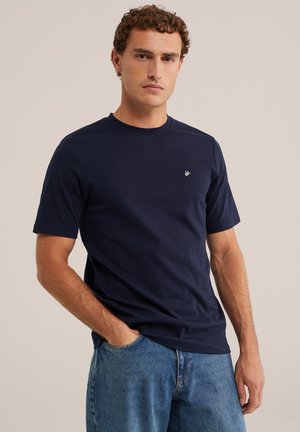 Jeune homme aux cheveux bouclés portant un t-shirt bleu marine à manches courtes et un jean bleu, debout avec une main dans la poche devant un fond neutre.