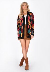 Donna che indossa un cardigan floreale colorato, una maglietta bianca, una mini gonna scamosciata marrone e stivali alla caviglia in suede marrone, in piedi su uno sfondo bianco.