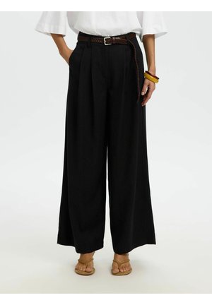 Personne portant un pantalon noir à taille haute et jambes larges avec une ceinture tressée marron, un haut blanc, des bracelets jaunes et rouges, et des sandales couleur cuir clair.