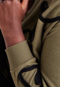 Oliwkowa zielona bluza z kapturem z czarnym ozdobnym obszyciem, ściągaczami na rękawach i teksturowaną tkaniną. Zbliżenie na szczegóły ramienia i dłoni.