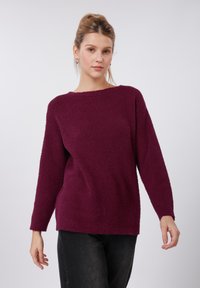 Pull bordeaux en tissu duveteux avec une coupe décontractée, un large col et des manches longues, associé à un jean foncé. Le tissu texturé apporte de la douceur.