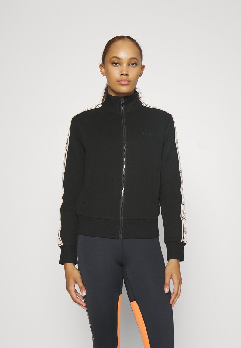 Veste à zip noire avec col montant, présentant un ruban contrastant sur les côtés et deux poches. Tissu doux avec des poignets et un ourlet côtelés.