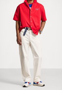 Red short-sleeve button-up shirt, white relaxed trousers, blue and red sneakers with black accents, and a striped belt. Features logo embroidery.