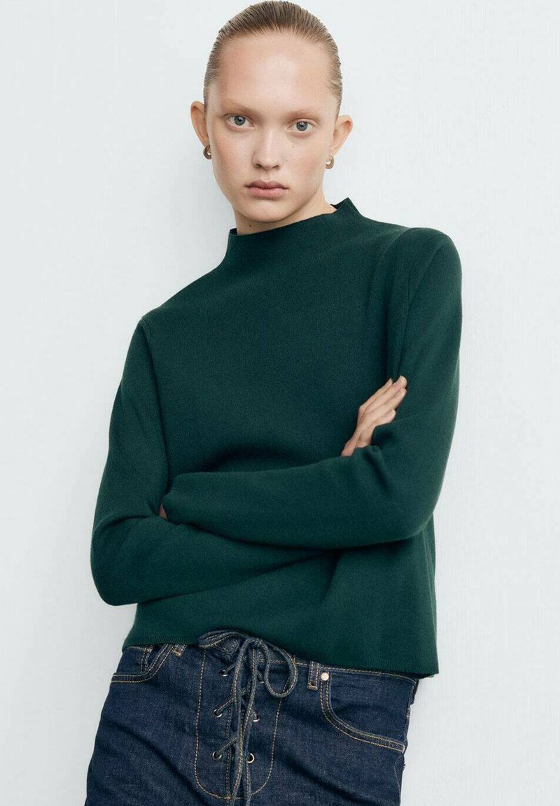 Maglione verde con collo alto, realizzato in tessuto morbido. Abbinato a jeans scuri, con parte anteriore con lacci e accessori in tonalità argento.