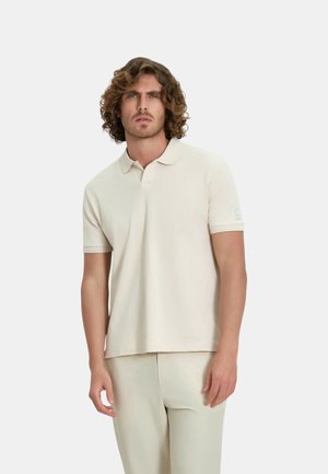Uomo con capelli ricci che indossa una polo beige chiaro a maniche corte e pantaloni abbinati, in piedi contro uno sfondo bianco semplice.