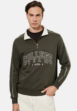 Młody mężczyzna ubrany w oliwkowo-zieloną bluzę rozpinaną do ćwierć długości z napisem "College Team" i białymi lamówkami na rękawach, w połączeniu z jasnymi spodniami.