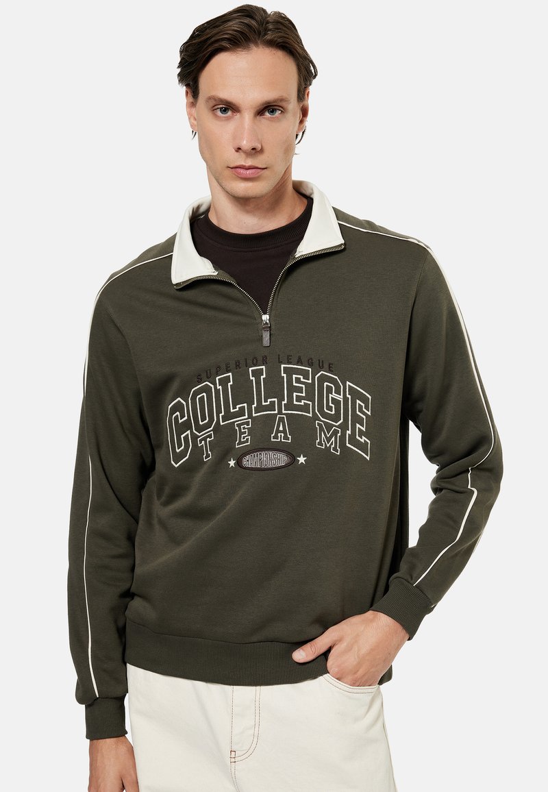 Jeune homme portant un sweat-shirt vert olive à fermeture éclair quart de zip avec le texte « College Team » et un liseré blanc sur les manches, associé à un pantalon clair.