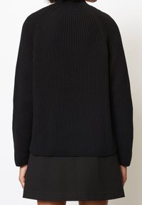 Pull noir en maille avec un col montant, une texture côtelée et de longues manches raglan. Porté sur une jupe noire, offrant une silhouette décontractée.
