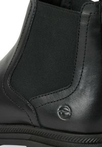 Botină din piele neagră, cu bandă elastică pe lateral, cusături decorative, textură netedă și un accent circular cu logo aproape de talpă.