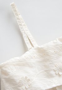 Haut en tissu blanc avec bretelles ajustables, orné d'une broderie florale et d'une surface texturée, mettant en valeur des détails de couture minutieux.