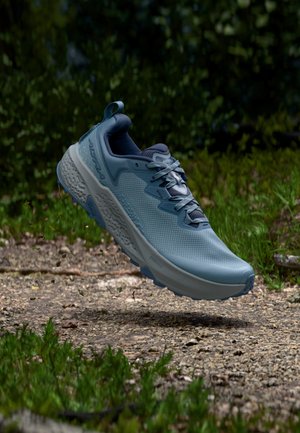 Hellblauer Trailrunning-Schuh mit dicker Sohle, der über einem Kiesweg schwebt, umgeben von grünem Laub.