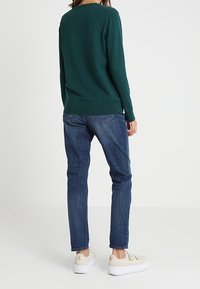 Vihreä neulepaita, siniset farkut ja beiget lenkkarit, joissa on teksturoitu pinta ja vaalea pohja. Näkymä takaa.