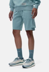 Sudadera de color azul claro con una etiqueta, combinada con pantalones cortos a juego de color azul claro que cuentan con un bolsillo lateral. Usada con zapatillas deportivas y calcetines de color azul claro.