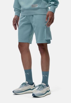 Sudadera de color azul claro con una etiqueta, combinada con pantalones cortos a juego de color azul claro que cuentan con un bolsillo lateral. Usada con zapatillas deportivas y calcetines de color azul claro.