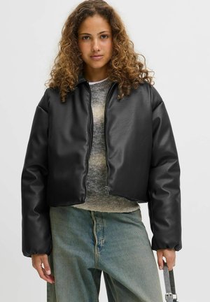 Sort cropped syntetisk læderjakke med en glat overflade, lynlås og afslappet pasform. Båret over en grå striktrøje og blå jeans.