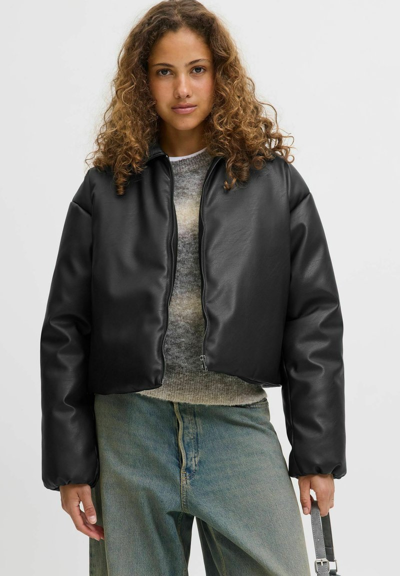 Czarna krótka kurtka z syntetycznej skóry o gładkiej teksturze, z zapięciem na zamek i luźnym kroju. Nosi ją osoba w szarej wełnianej bluzie i niebieskich dżinsach.