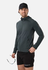 Moški v temni jopiči z zadrgo in kapuco ter črnih kratkih hlačah, ki v eni roki drži športna sončna očala in rahlo gleda v desno.