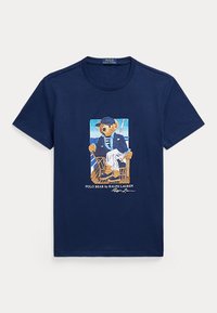 T-shirt bleu marine avec un motif d'un ours en tenue nautique assis sur une chaise, avec le texte "Polo Bear by Ralph Lauren" en dessous.