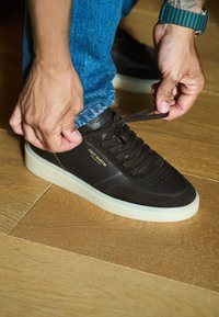 Czarny sneaker wykonany z zamszu i skóry, z teksturowaną powierzchnią, płaskimi brązowymi sznurowadłami oraz jasną podeszwą. Widoczne logo "FRED MARTIN".