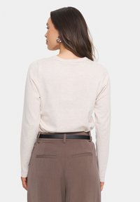 Svetlo bež pulover z dolgimi rokavi in okroglo izrezom, v kombinaciji z rjavimi hlačami visokega pasu, ki imajo črn pas in zadnje žepke.
