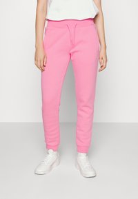 Pantalones de chándal rosa con cintura ajustable, puños entallados y bolsillos laterales, hechos de una tela suave y texturizada. Combinados con zapatillas blancas.