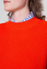 Jasnopomarańczowy, dziany sweter o ribbowej strukturze i wysokim kołnierzu, nałożony na niebieską koszulę z kołnierzykiem w białe abstrakcyjne wzory.