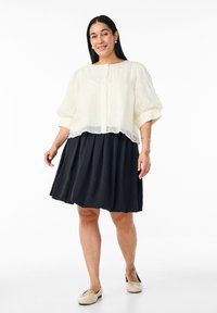 Bílý texturovaný cropped top s nafouknutými rukávy, vrstvený nad černou skládací sukní ke kolenům. Na nohou jsou světle zbarvené baleríny.