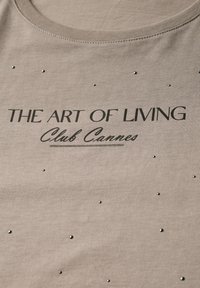 Šedé tričko z látky s potiskem "UMĚNÍ ŽÍT Club Cannes" v černé barvě, zdobené malými dekorativními cvočky rovnoměrně rozmístěnými.