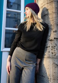 Maglione nero a maniche lunghe, abbinato a pantaloni grigi con trama, davanti a un muro di pietra. Il modello indossa un berretto lavorato a maglia colore viola scuro.