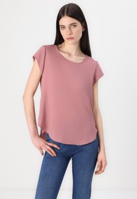 Top rosa pallido a maniche corte con scollatura rotonda e orlo arrotondato, realizzato in tessuto leggero, abbinato a jeans blu aderenti.