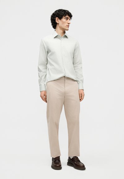 Jeune homme aux cheveux noirs bouclés portant une chemise à manches longues à rayures claires, un pantalon beige et des chaussures marron foncé à lacets, debout et regardant vers la droite.