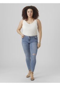 White ribbed tank top, blue ripped skinny jeans, and black strappy sandals. Hair is curly and voluminous, with hoop earrings.