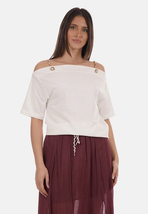 Donna che indossa una maglia bianca scollata con spalline a catena dorata e una gonna con coulisse color bordeaux, in piedi davanti a uno sfondo semplice.