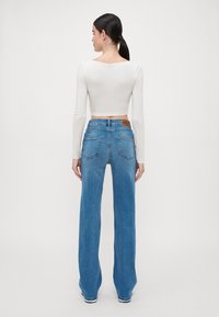 Helle, langärmlige, kurze Bluse kombiniert mit blauen weiten Jeans. Die Jeans haben Gesäßtaschen und ein geprägtes Lederpatch an der Taille.