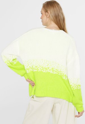 Frau trägt einen weißen und leuchtend grünen gestrickten Pullover mit Farbverlaufsmuster, steht mit einer Hand in der Hüfte und ist mit dem Rücken zur Kamera gewandt.