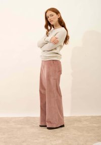 Jasnoszary sweter z teksturą i długimi rękawami, w połączeniu z różowymi szerokimi spodniami z korduroyu i czarnymi butami na obcasie. Proste tło.