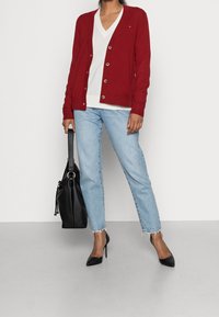 Červený pletený kardigan s knoflíkovým zapínáním, bílé tričko s výstřihem do V, světle modré džíny se rovnými nohavicemi, černá kabelka a černé střevíce na podpatku.