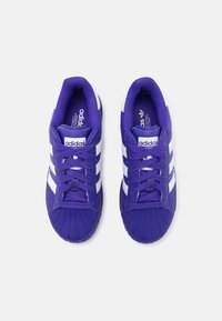 Ett par lila Adidas sneakers med vita ränder sedd ovanifrån, med skalkälsdesign och snörning på en vit bakgrund.