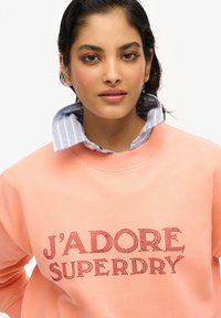 Sudadera color durazno con "J'ADORE SUPERDRY" en brillantes rhinestones rojos. Llevada sobre una camisa de cuello azul claro y blanco a rayas.