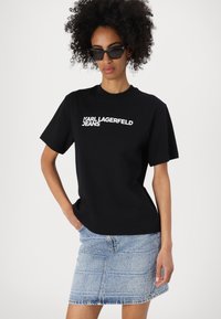 Črna bombažna majica s kratkimi rokavi in belim napisom "KARL LAGERFELD JEANS", v kombinaciji s svetlo modrim jeans krilom.