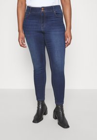 Mørkeblå slim-fit jeans med to bronsknapper i høj talje, der har subtil falming og syede sømme, båret med sorte stilethæle.