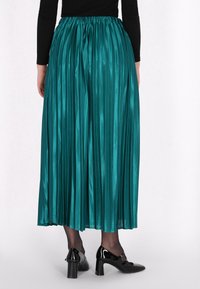 Teal selyem anyagú, pliszírozott szoknya, rugalmas derékkal, amely a vádli közepéig ér, fekete lakkbőr magassarkú cipőkkel párosítva.