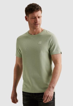 Čovjek gleda dolje noseći svijetlozelenu kratkih rukava pletenu majicu i tamne hlače na jednobojnoj sivoj pozadini.