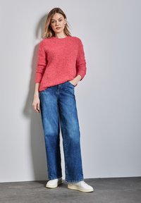 Różowy puszysty sweter z okrągłym dekoltem, zestawiony z szerokimi niebieskimi dżinsami i białymi trampkami, na jednolitym tle.